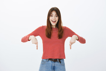 Incredible place for advertisement. Portrait of overwhelmed fascinated young caucasian woman with positive friendly attitude, being charmed with exciting news, pointing down over gray background