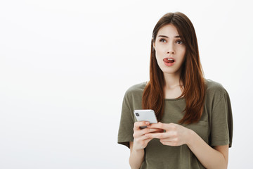 Fototapeta premium Pretty thoughtful female student holding smartphone, looking up and sticking out tongue while thinking or counting in mind, making grocery list and typing items in phone to remember