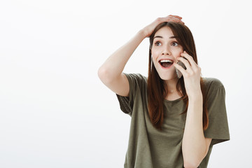 Woman plesantly shocked being happy for friend who won in lottery. Excited joyful young female in casual clothes, holding hand on head, smiling broadly while talking on smartphone over gray wall