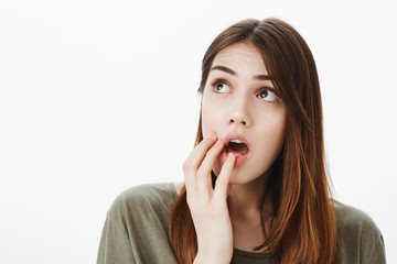 Portrait of confused unfocused good-looking caucasian woman, standing with opened mouth over gray background, holding finger on lip and looking up, dreaming during office meeting