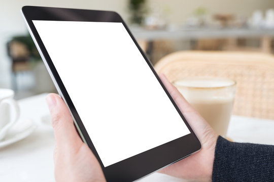 Mockup image of hands holding black tablet pc with blank white screen and coffee cup on table in cafe