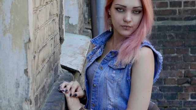 Teenage girl with nose and lips piercing and pink hair standing on brick wall background. Slow motion. HD
