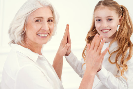 Never get bored. Radiant grandmother and her granddaughter looking into the camera with a smile on their faces while having fun and playing clapping game together. - Powered by Adobe