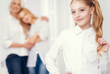 Fototapeta premium Children are our future. Selective focus on a cheerful little lady touching her ponytail and lightly smiling into the camera while her tender mother and grandmother hugging with love behind.