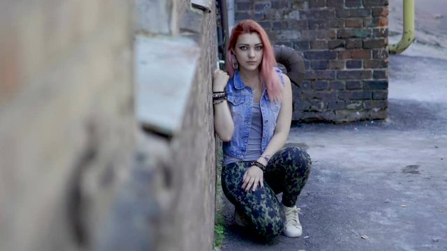 Teenage girl with nose and lips piercing and pink hair sit leaning against brick wall and looking at camera. Slow motion. HD