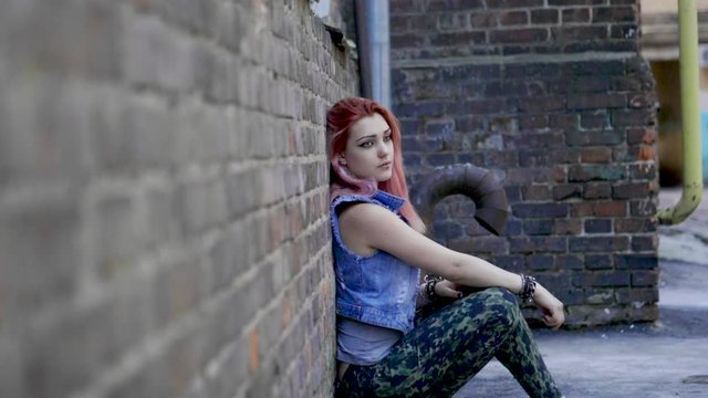 Teenage girl with nose and lips piercing and pink hair sit leaning against brick wall. Slow motion. HD
