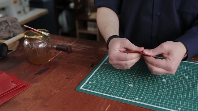 Leathersmith is applying glue on a leather parts of the zipper with a special brush. He works at the table in the workshop.