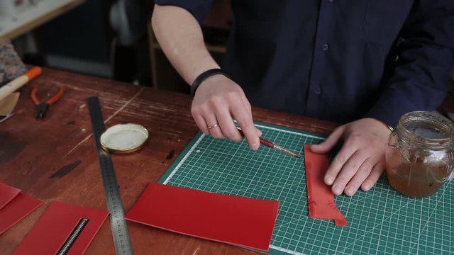 Leathersmith is applying glue on a leather parts of the zipper with a special brush. He works at the table in the workshop.