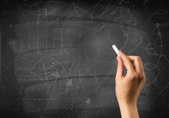 Female hand holding white chalk in front of a blackboard with scribbles and plans drawn on it 