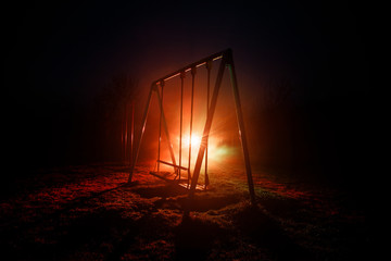 Night photo of metal swing standing outdoor at night time with fog and surreal toned light on background. Nobody there.