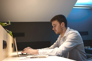 Sideways portrait of focused young male office worker typing on portable computer, filling in...