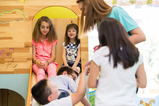 Cute pre-school girls smiling while sitting on a wooden slide during playtime supervised by a young teacher in the classroom of a modern kindergarten