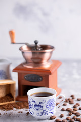 Grains of coffee fall out of a vintage coffee grinder. Hot black coffee in a beautiful porcelain cup on the table. A beautiful coffee composition with a place under your text.