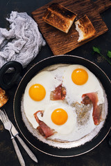 Fried eggs in a frying pan with bacon, toast, napkin, forks. Scrambled egg with bacon on a dark background, top view.