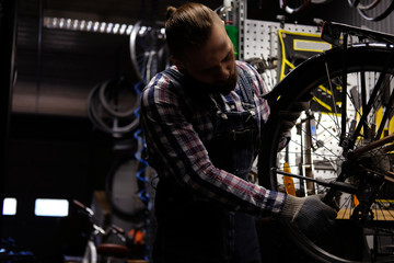 Naklejka na ściany i meble Handsome stylish male wearing a flannel shirt and jeans coverall, working with a bicycle wheel in a repair shop. A worker using a wrench mounts the wheel on a bike in a workshop.