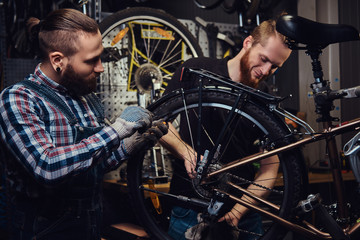 Obraz na płótnie Canvas Two handsome stylish male working with a bicycle in a repair shop. Workers repair and mounts bike in a workshop
