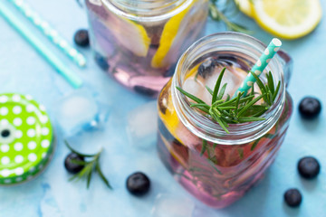 Lemonade or cocktail with blueberries and rosemary, cold refreshing drink with ice on a blue concrete or stone table.