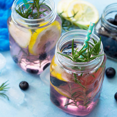 Lemonade or cocktail with blueberries and rosemary, cold refreshing drink with ice on a blue concrete or stone table.