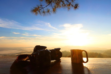 DSLR camera and coffee glass on wood table have view of fog, mountain, cloud, blue sky and flare orange color light of sunrise is background at Khao Kho Royal Palace, Phetchabun,Thailand