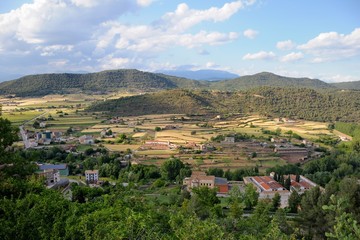 Fototapeta na wymiar View from Cardona Castle. Sunlit large valley with colorful fields and small houses. The valley is surrounded by green mountains. Blue sky and white clouds. Summer. Catalonia, Spain.