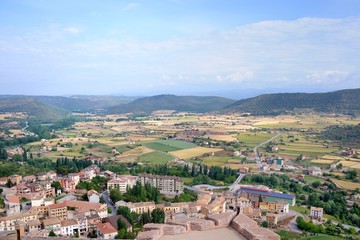 Fototapeta premium View from Cardona Castle. Sunlit large valley with colorful fields and small houses. The valley is surrounded by green mountains. Blue sky and white clouds. Summer. Catalonia, Spain.