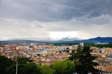 View of the city of Girona from the medieval pedestrian border wall. Roofs of houses, trees. Storm clouds over the city, somewhere sunlight makes its way through the clouds. Girona, Spain