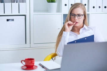 Young beauiful Caucasian businesswoman in glasses intently working on notebook computer and business document at office.