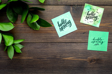 Spring lettering, spring motto. Lettering hello spring on stickers among green foliages on dark wooden table top view copy space