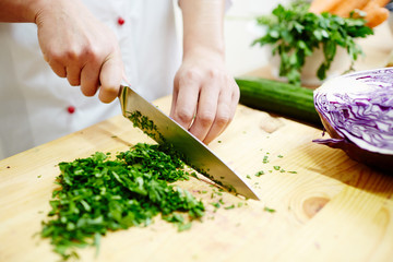 Human hands with sharp steel knife shredding green parsley leaves on wooden board for salad or stew