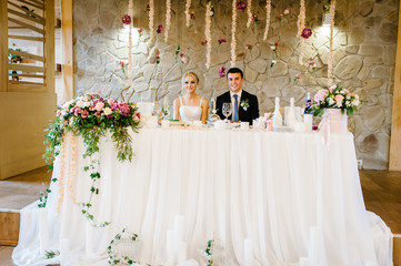 The bride and groom sit at the table and celebrate the wedding in the banquet hall of the restaurant. Newlyweds of the indoors. Wedding ceremony decorated with flowers and greenery.