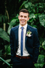 Happy handsome groom in a suit and tie is standing on the background of greenery, on the jacket is...