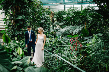 Portrait the groom in wedding suit and the bride in dress are going in the Botanical green garden...