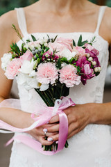 Obraz na płótnie Canvas Young beautiful bride in an elegant dress is standing and holding bouquet of pink and purple flowers and greens with ribbon at the wedding ceremony.