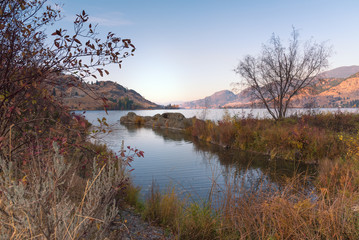 Obraz premium Landscape of calm lake with autumn leaves and grasses in foreground and sunset light on distant mountains.