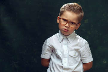 Serious little boy pupil of primary classes with glasses and white shirt angrily looks into the camera and hides his hands behind his back on dark background.