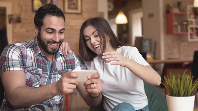 Cute couple playing game on modern smartphone, pressing on gadget screen with great enthusiasm, black-haired man and attractive brunette enjoying leisure time in warm european cafe, expressing