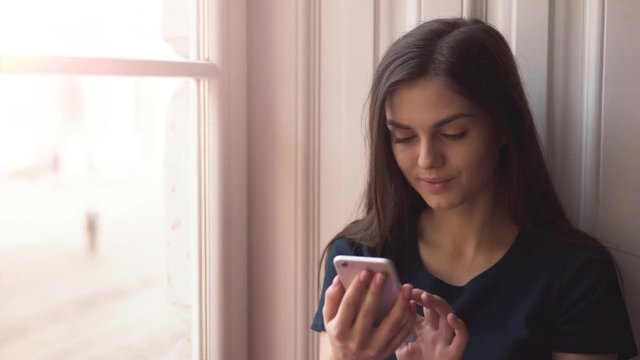 Beautiful dreaming woman typing a message near the window, using the gadget with great pleasure, inloved girl chatting on phone in cozy apartment on ordinary weekday