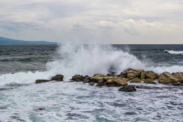 Powerful wave in sea crashing against rock sending up sprays of white foam in sky with cirrus clouds seagull above horizon line
