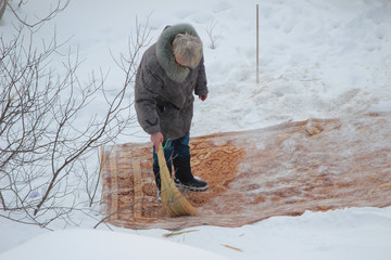 An elderly woman cleans the carpet in the snow in winter