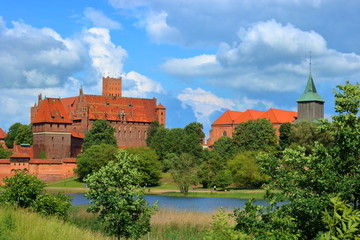 Fototapeta na wymiar Malowniczy widok zamku krzyżackiego w Malborku, Polska, z przeciwnego brzegu rzeki Nugat, z malowniczymi obłokami na błękitnym niebie