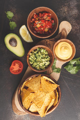 Mexican food concept. Nachos - yellow corn totopos chips with various sauces in wooden bowls: guacamole, cheese sauce, pico del gallo. Top view, food background