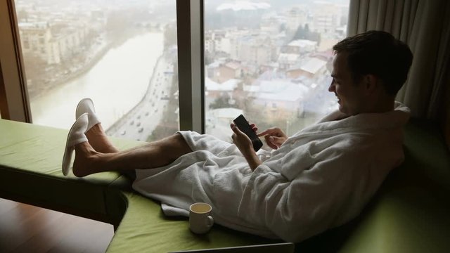 Young smiling handsome man wearing bathrobe messaging on a mobile phone and talking. He is sitting near window with great city view with cup of coffee at luxury scyscraper hotel.