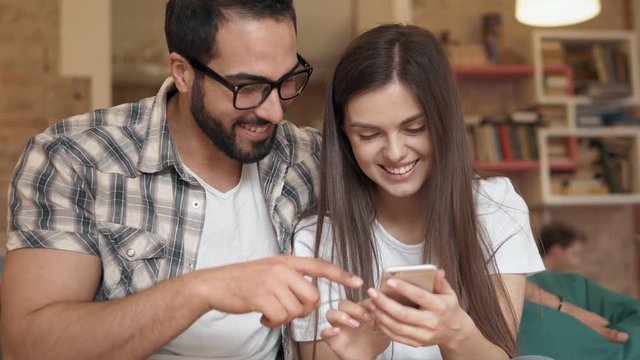 Couple selecting best selfies on phone, intelligent bearded man in glasses pointing at gadget screen, cute slender woman holding smartphone cheerfully, sitting on green comfortable chair in cozy cafe