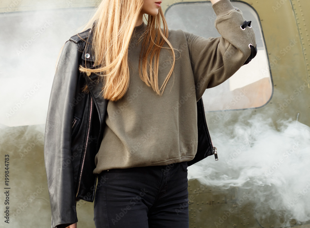 Wall mural Beautiful fashion young woman in a black leather jacket standing at the old airfield shed in summer in the clubs of smoke