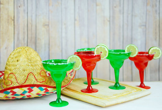 Margaritas In Red And Green Glasses With Salted Rims And Lime Garnishes Sitting On A Wooden Table With A Sombrero For The Mexican Holiday Cinco De Mayo