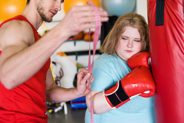 Waist up portrait of exhausted obese woman looking at measuring tape during training in gym