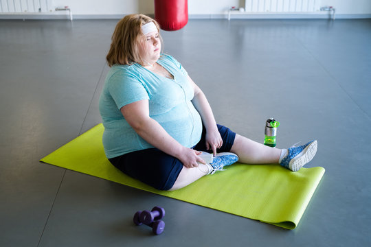 Full Length Portrait Of Tired Obese Woman Sitting On Yoga Mat And Resting After Fitness Training In Gym