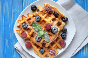Traditional belgian waffles with fresh berrys and sugar powder on white plate, blue wooden background. Flat lay, top view, copy space.