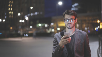 A young man is holding scrolling texting in his cellphone. A serious man calls for a taxi in an app in night time