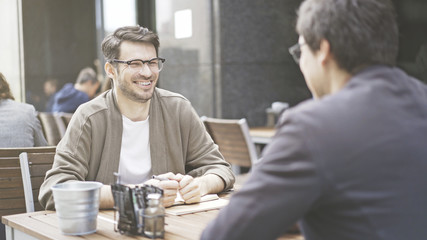 Two friends are laughing having a conversation at table of the cafe outdoors. A man dressed in a jacket wearing eyeglasses holding a pen